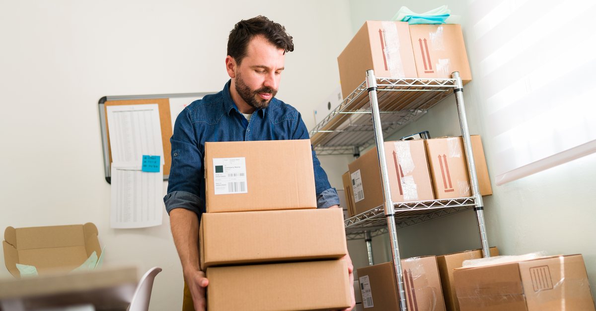 Online shop owner carrying boxes beside shelves of parcels